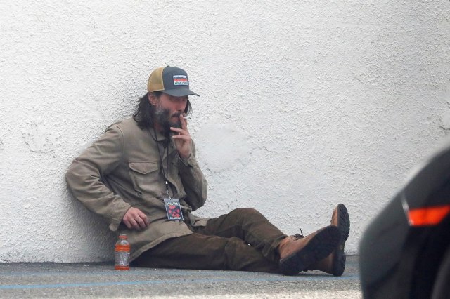 Киану Ривс како бездомник со седа брада и долга коса пуши цигара пред клуб